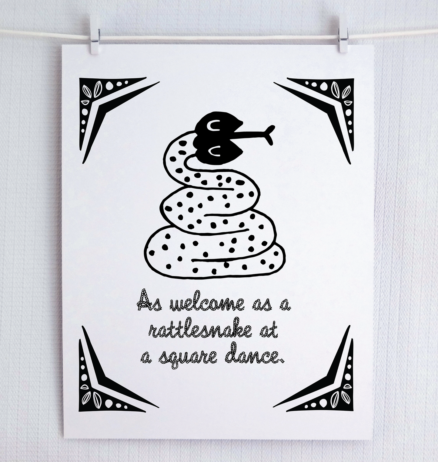 An illustrated rattle snake with friendly eyes and polka dots sits over a rope-style font that says “As welcome as a rattlesnake at a square dance.” Decorative corner design reminiscent of the wild west.
