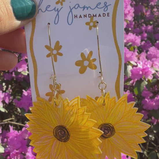 Yellow sunflower earrings on a card with a blurred background of pink flowers