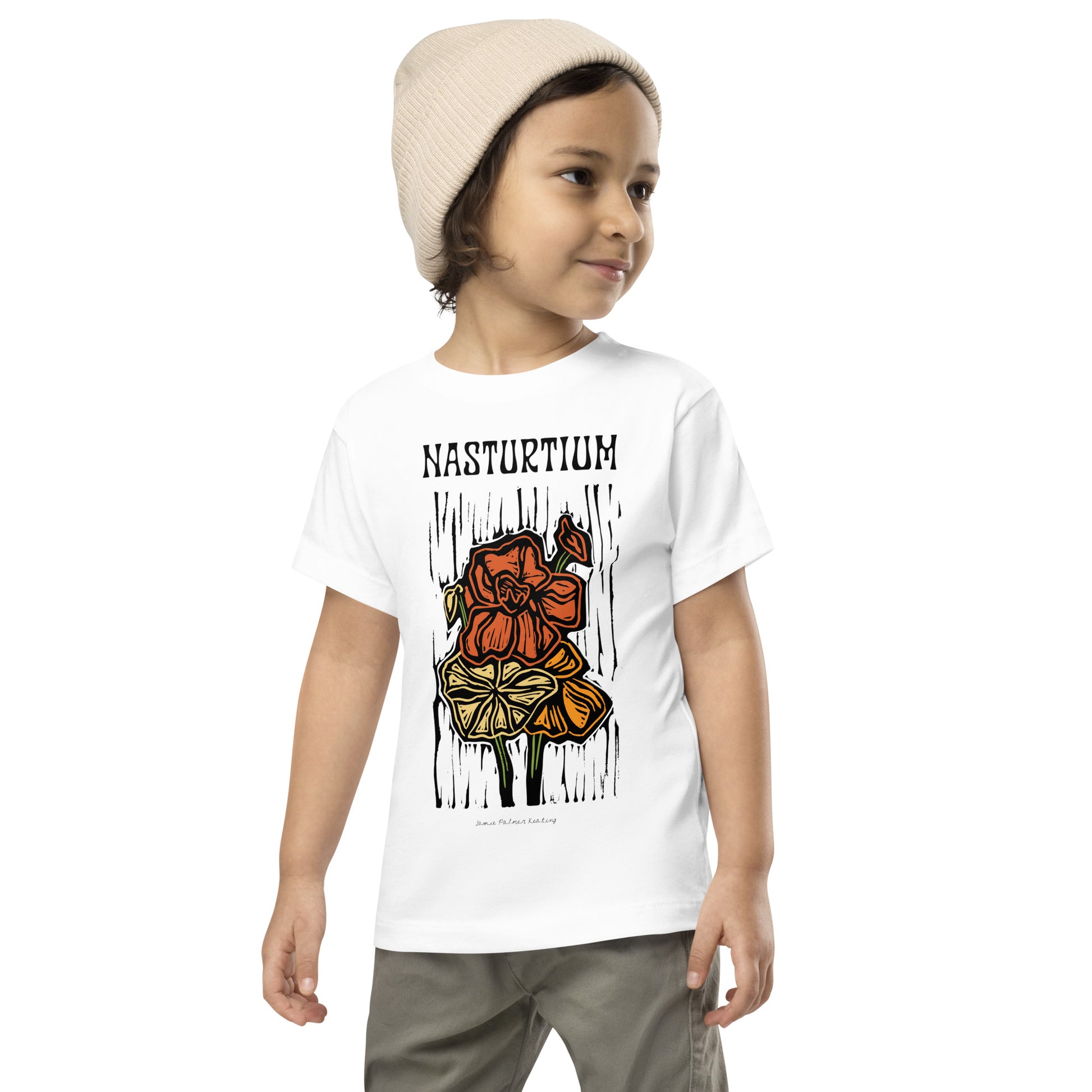 A child wearing a white short-sleeve graphic T-shirt with the word 'NASTURIUM' and a stylized flower illustration on the front, paired with gray pants and a beige knit beanie, against a plain white background.