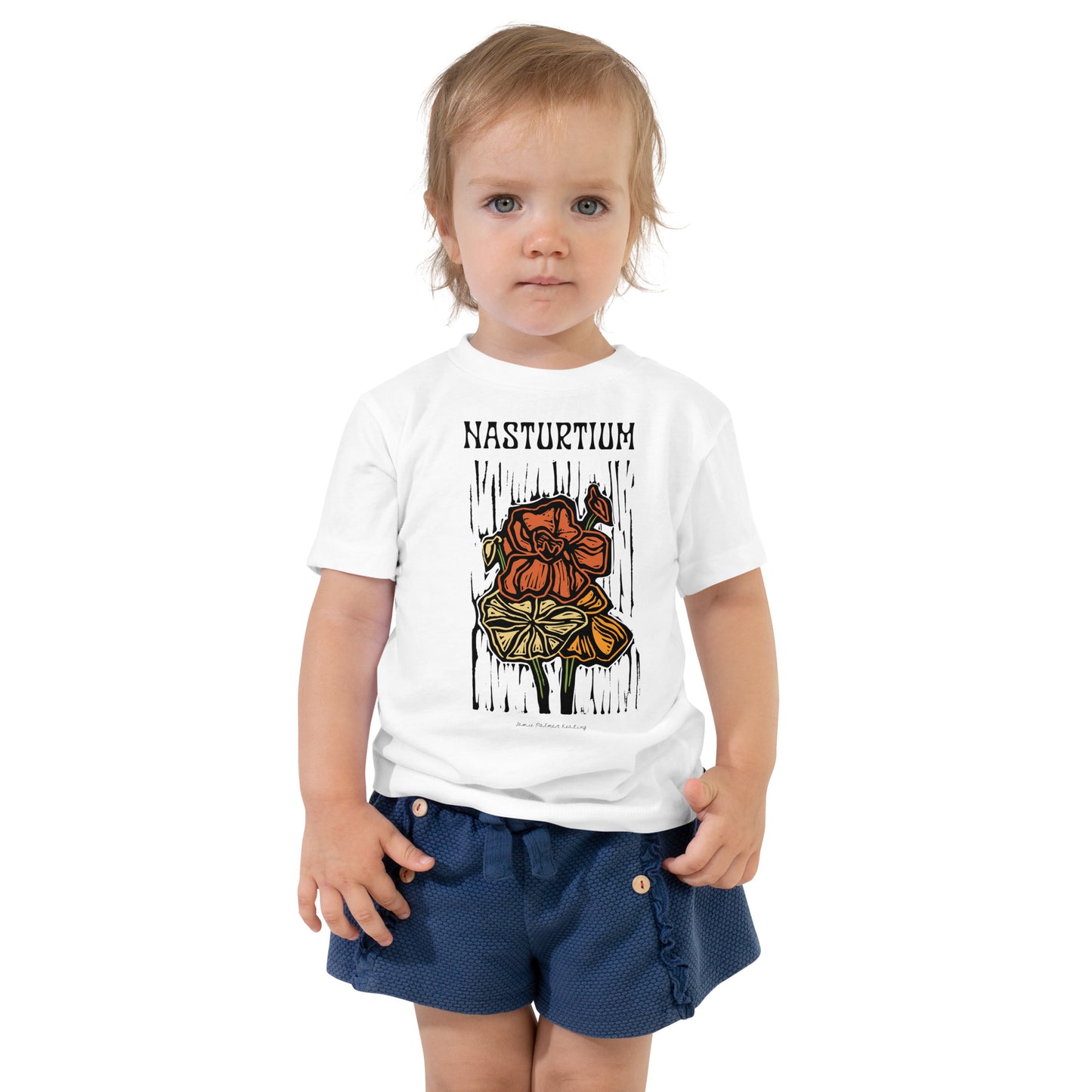 A toddler standing against a white background wearing a white graphic t-shirt with 'NASTURTIUM' and a flower illustration, paired with navy shorts.