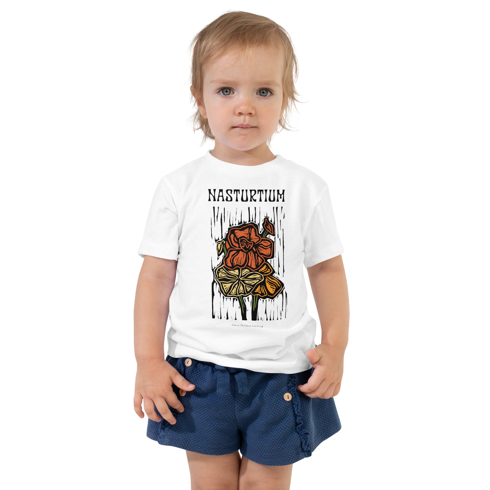 A toddler standing against a white background wearing a white graphic t-shirt with 'NASTURTIUM' and a flower illustration, paired with navy shorts.