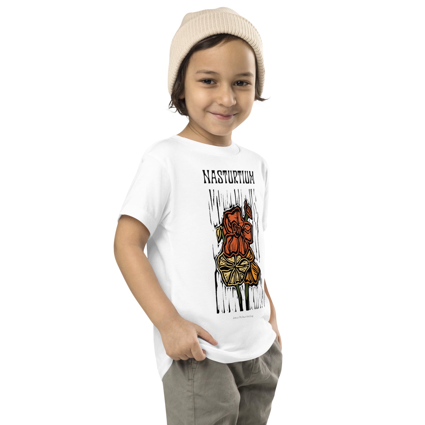 A young child wearing a white short-sleeve graphic T-shirt and a beige knit beanie, standing against a plain white background. The shirt features the word 'NASTURTIUM' above a stylized orange and yellow figure with black vertical lines.