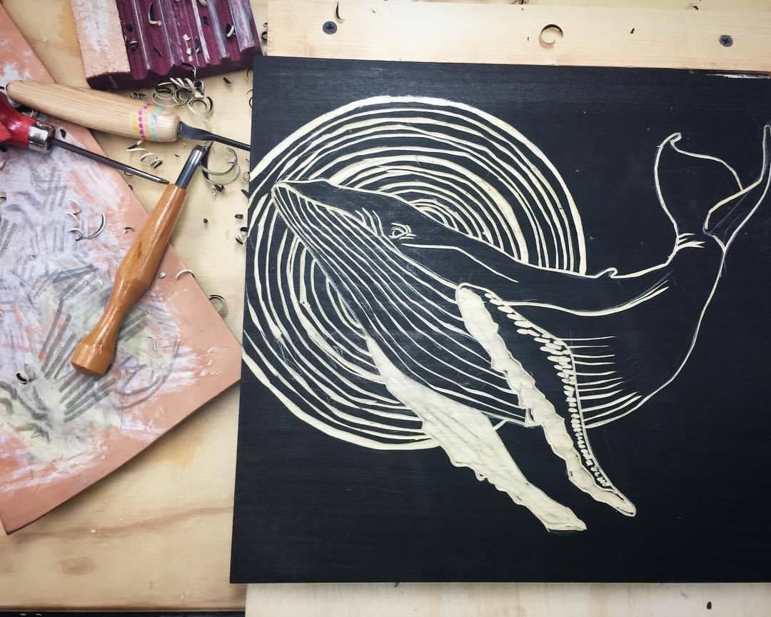 Woodcut print of a whale on black wood with printing tools in the background