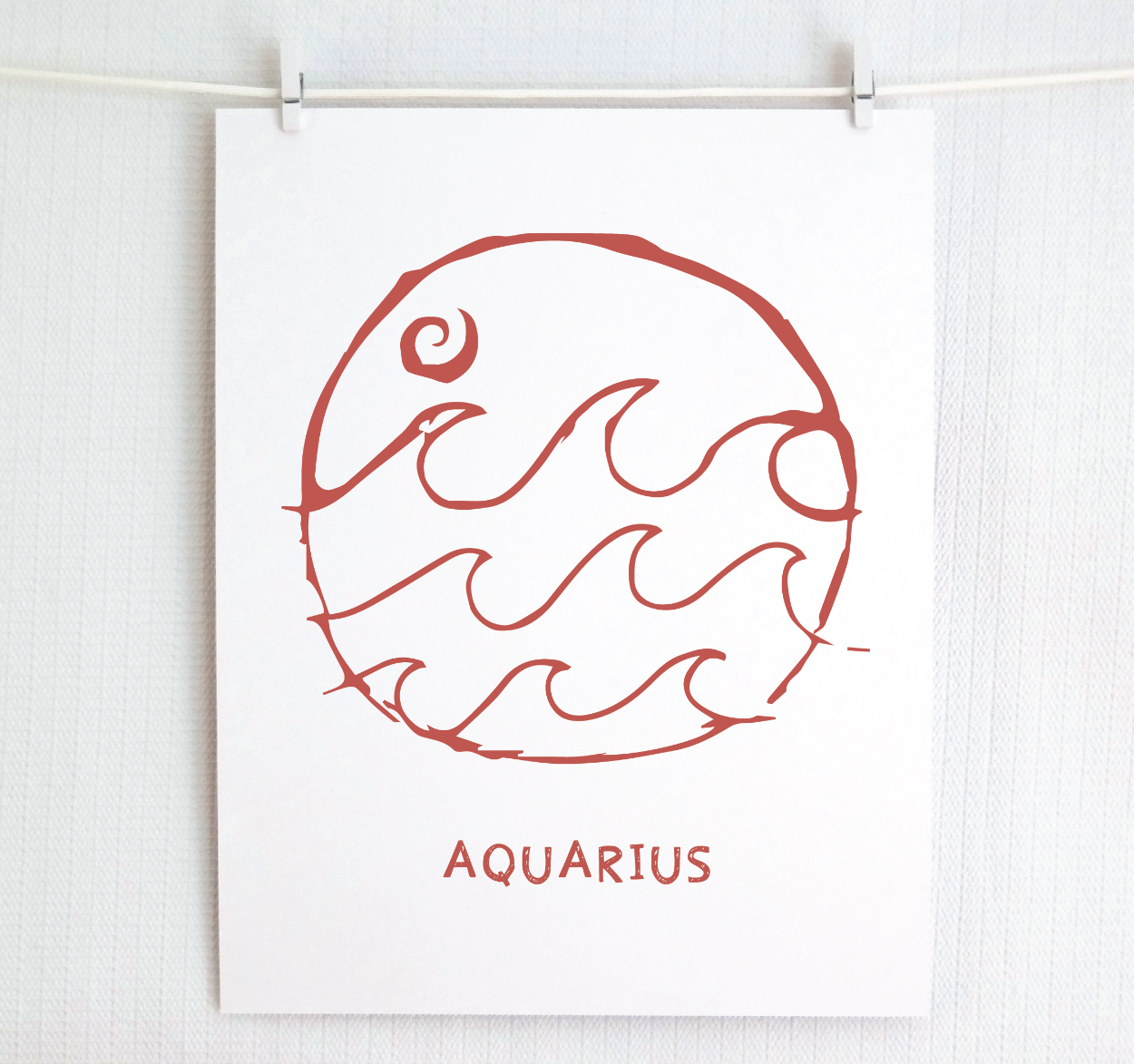 An illustration of the Aquarius Zodiac sign, printed on paper with the word 'AQUARIUS' at the bottom in blush