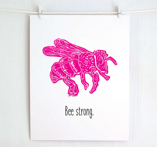 A digital print of a linoleum block illustration depicting a pink bee with the phrase 'Bee strong' underneath it, displayed on a white background.