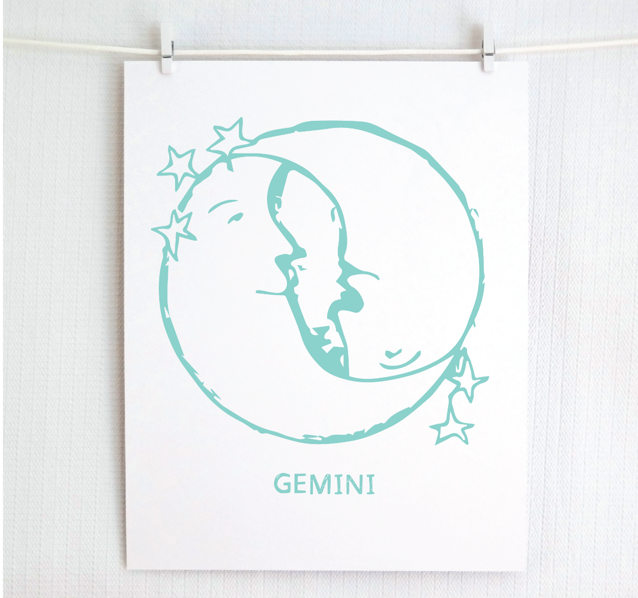 An illustration of the GEMINI Zodiac sign as crescent moons, printed on paper with the word 'GEMINI’ at the bottom in light teal