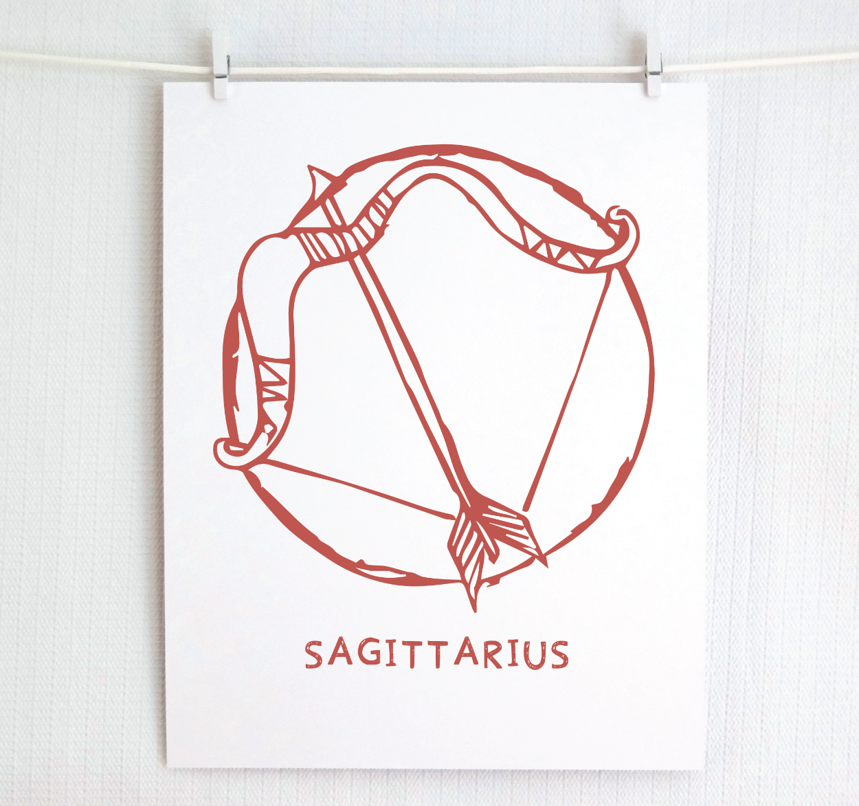 An illustration of the SAGITTARIUS Zodiac sign as a bow and arrow, printed on paper with the word ‘SAGITTARIUS’ at the bottom in blush.