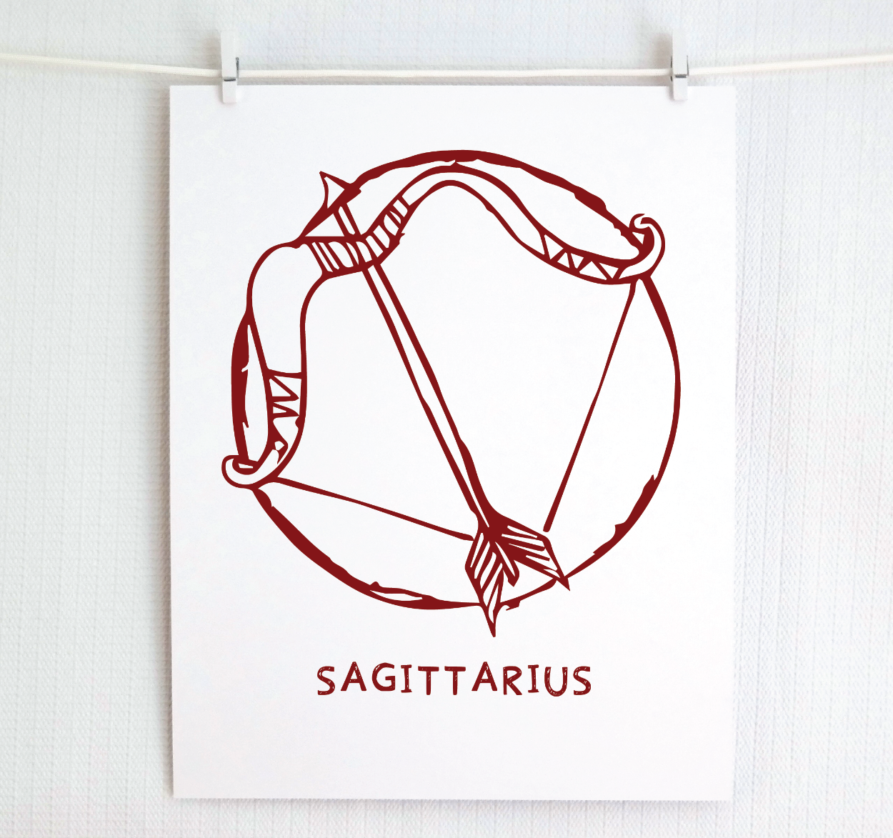 An illustration of the SAGITTARIUS Zodiac sign as a bow and arrow, printed on paper with the word ‘SAGITTARIUS’ at the bottom in cranberry.